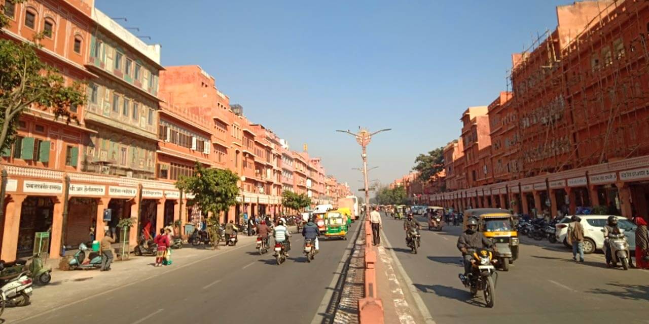 Front view of Johari Bazaar, a bustling market street in Jaipur, Rajasthan, India. The road is lined with colorful shops and vibrant awnings, with pedestrians and vehicles navigating through the busy thoroughfare. People are seen browsing through the various stalls and shops selling traditional Rajasthani jewelry, textiles, and other handicrafts.