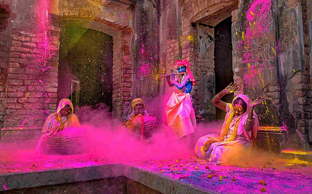 Lord Krishna playing the flute while seated atop a colorful platform, surrounded by vibrant hues of gulal (colored powder), as devotees play the dholak drum below during the festive celebration of Holi.