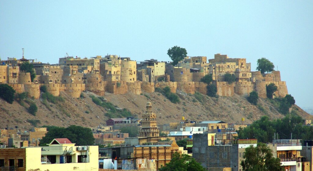 Aerial view of the majestic Jaisalmer Fort rising above the golden desert landscape, showcasing its intricate sandstone architecture and fortified walls under the blue sky.