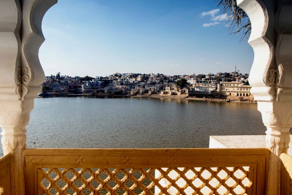 A tranquil scene of Pushkar Lake viewed through a large window, with the soft glow of sunlight illuminating the surroundings.
