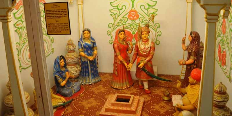 Image sourced from Google showing a traditional Indian wedding ceremony with a bride, groom, and priest performing rituals around a sacred fire in a mandap. Clay figurines of women are also present.