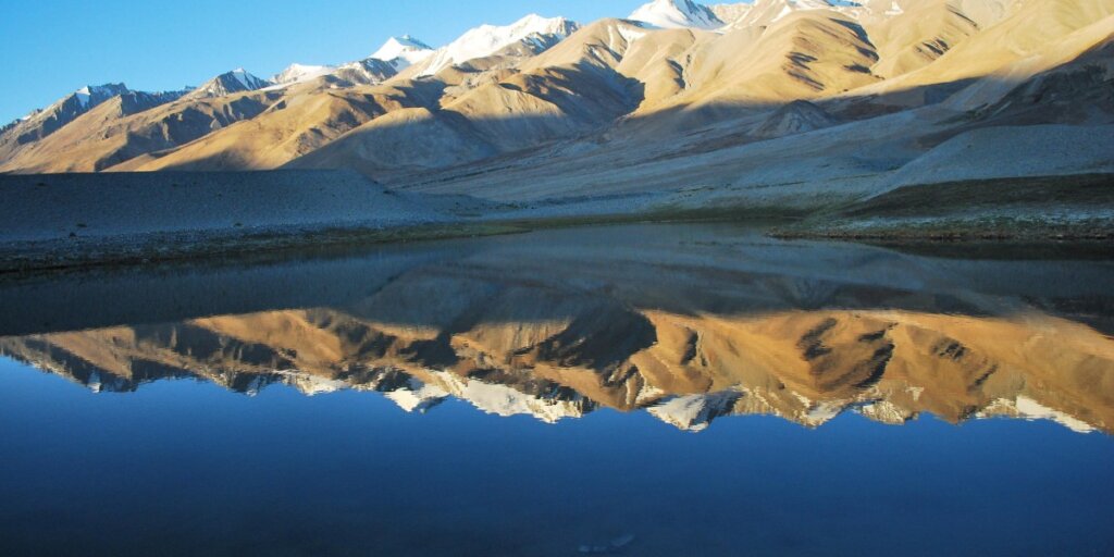 Reflective waters of Pangong Lake in Ladakh under a clear blue sky, showcasing the serene and vast landscape typical of this high-altitude region. Image by Wikimedia Commons.