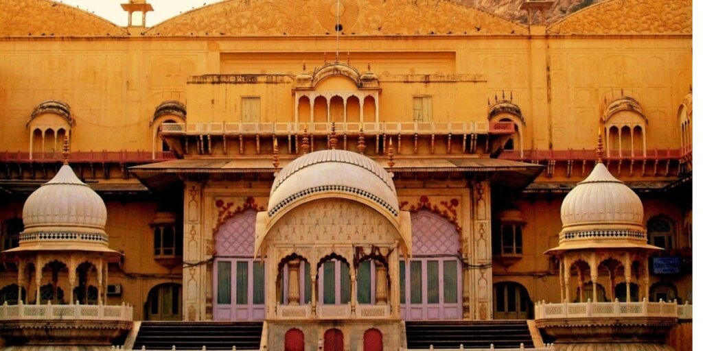 Front view of Alwar City Palace in Rajasthan, India. The facade features a central dome flanked by two smaller domes, intricate carvings, arched windows, and yellow-hued walls with decorative elements.