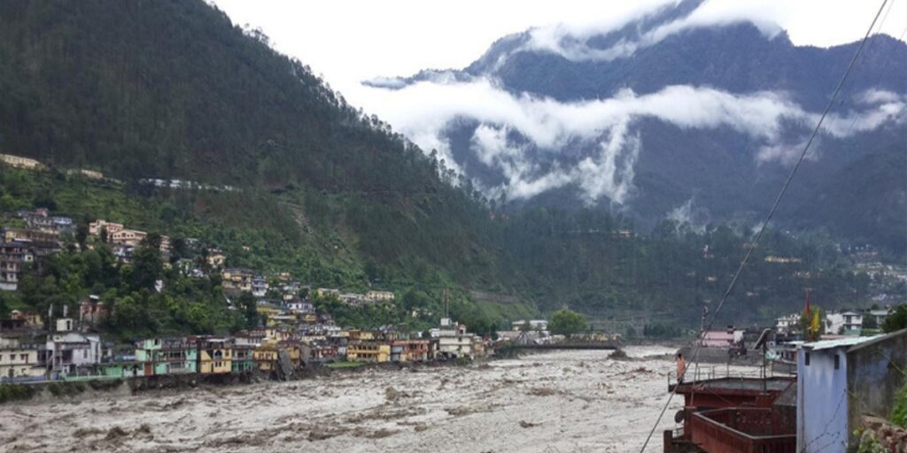 text: "A serene riverside scene in Barkot, Uttarakhand, India, featuring houses nestled among trees along the banks of a flowing river. The image captures the tranquil ambiance of this riverside settlement amidst the natural beauty of Uttarakhand. 