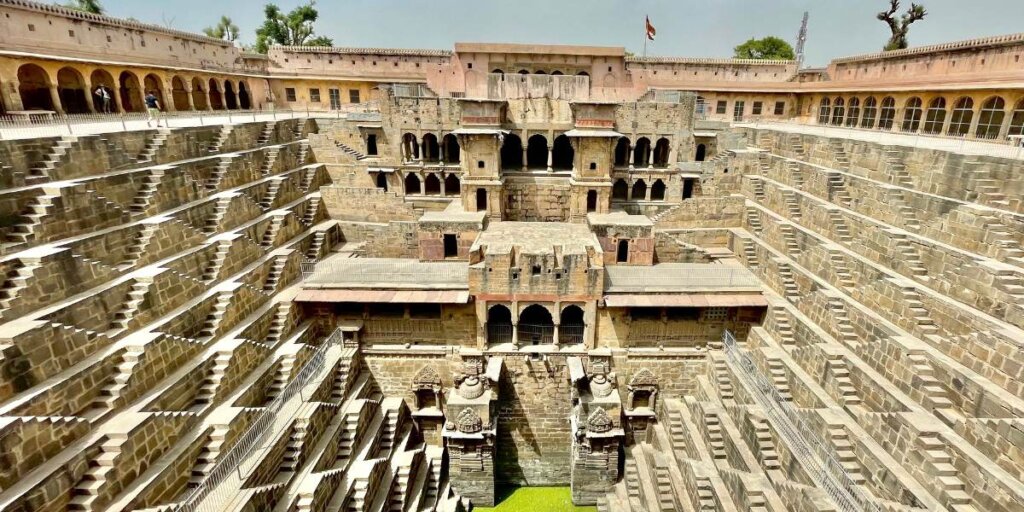 View of Chand Baori, a stepwell in Abhaneri, Rajasthan, India, showcasing its intricate geometric design. The stepwell features symmetrical staircases on three sides leading down to the water, with a multi-storied pavilion and ornate arches on the fourth side. The stone architecture displays detailed carvings and the well is surrounded by high walls.