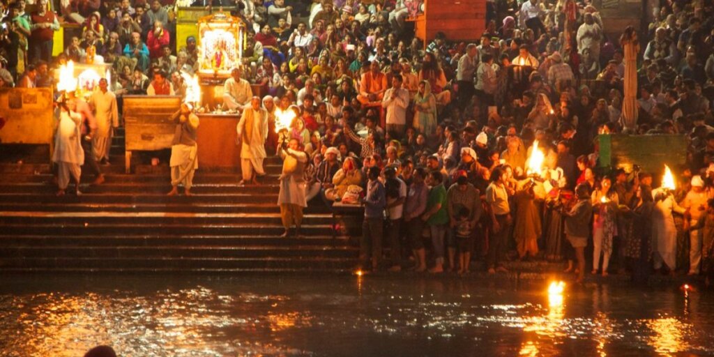 A close-up view of the Ganga Aarti ceremony in Haridwar, India, showing priests conducting rituals by the riverbank at dusk. Devotees gather around, and the flickering flames of ceremonial lamps illuminate the scene against the evening sky, evoking a sense of reverence and spirituality. Image by Flickr."