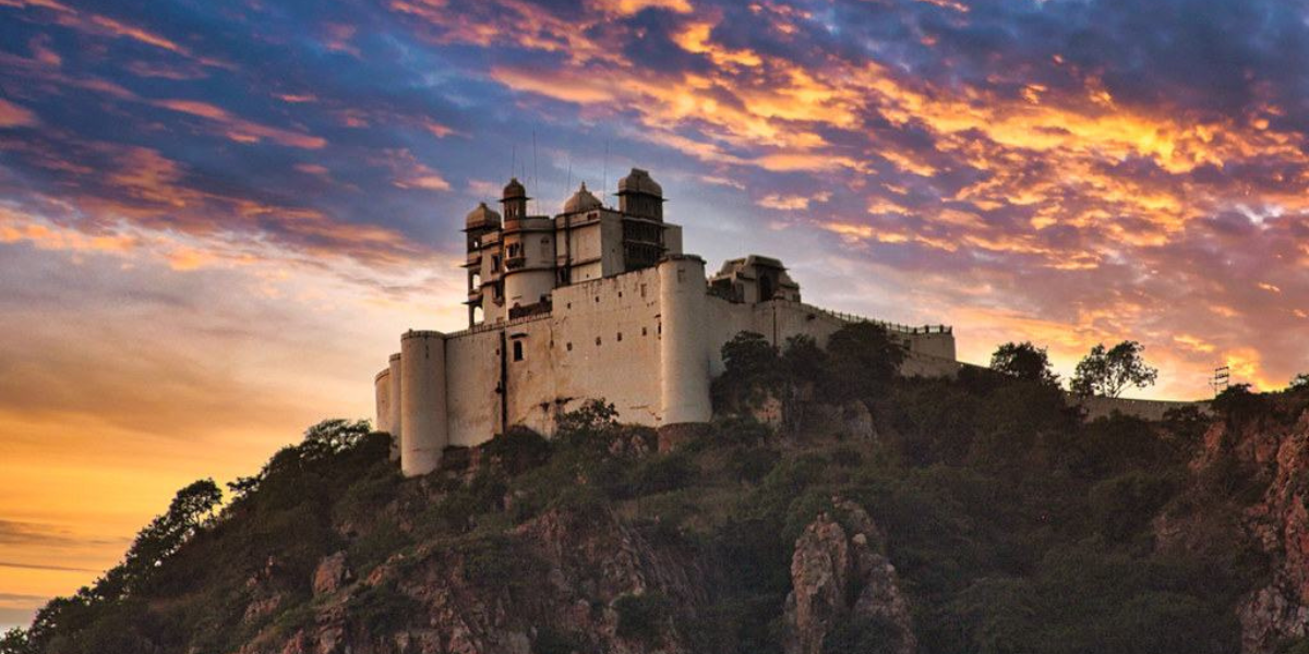  A dramatic view of the Sajjangarh Palace, also known as the Monsoon Palace, in Udaipur, Rajasthan, perched atop a hill against a vibrant, colorful sunset sky. The palace's white walls and architectural details are highlighted by the warm hues of the setting sun, creating a striking contrast with the surrounding greenery and rugged terrain.