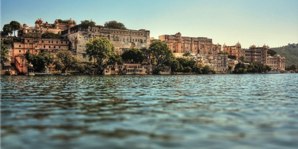 A stunning view of the City Palace complex in Udaipur, Rajasthan, seen from the waters of Lake Pichola on a clear day. The historic architecture of the palace buildings rises majestically from the lakeside, surrounded by lush greenery and hills in the background. (Image credit: Flickr)