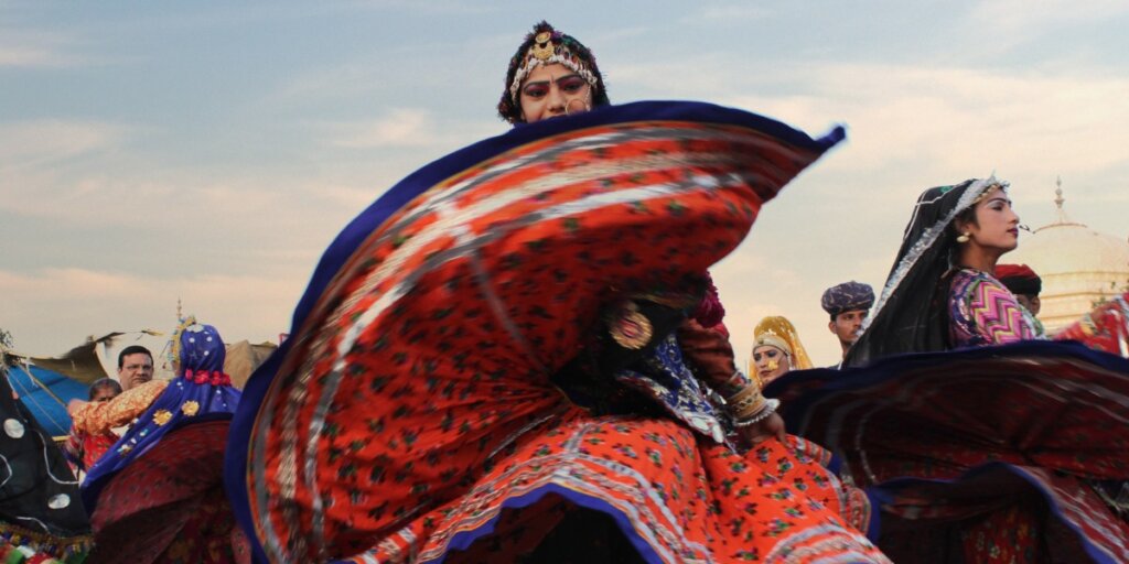  A group of Rajasthani dancers, dressed in vibrant traditional attire, perform a folk dance. The women are wearing colorful ghagras (skirts), cholis (blouses), and odhnis (scarves) adorned with intricate embroidery and mirror work. The men are in equally colorful kurtas and turbans. The dancers' movements are graceful and synchronized, reflecting the rich cultural heritage of Rajasthan.

Credit: Wikimedia Commons