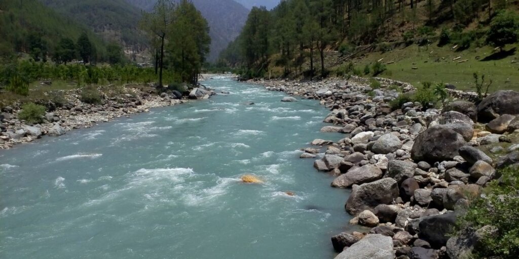 A scenic view of Uttarkashi, Uttarakhand, India, with a river flowing amidst lush greenery, surrounded by verdant hills. Image by Wikimedia Commons.