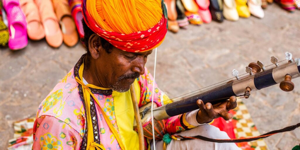 A man in traditional Rajasthani clothing is playing a musical instrument. He is surrounded by the vibrant atmosphere of Rajasthan, with colorful decorations and lively surroundings. 

Get this image on: Pexels | License details
Creator: Swastik Arora 
