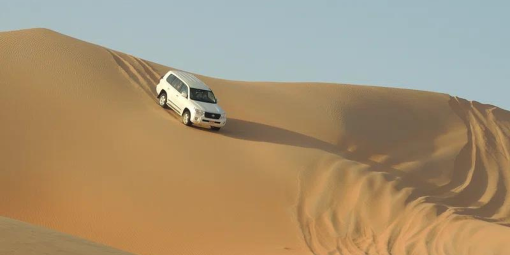 An SUV descends a sand dune during a desert safari. The vehicle kicks up a cloud of sand as it travels down a steep slope, showing the thrill of off-road desert driving. The vast desert landscape stretches in the background, with undulating dunes under a clear blue sky. This image shows the thrill and beauty of a desert safari. Get this image on: Rawpixel 