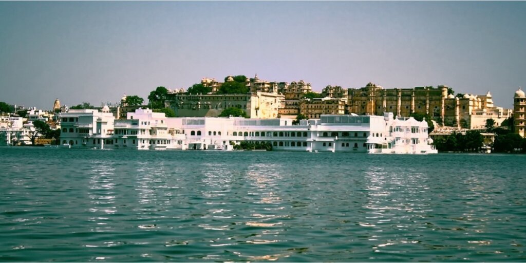  A serene view of Lake Pichola in Udaipur, surrounded by historic buildings and lush greenery, with calm waters reflecting the scenery.Image credit: Daniel Mennerich via Flickr