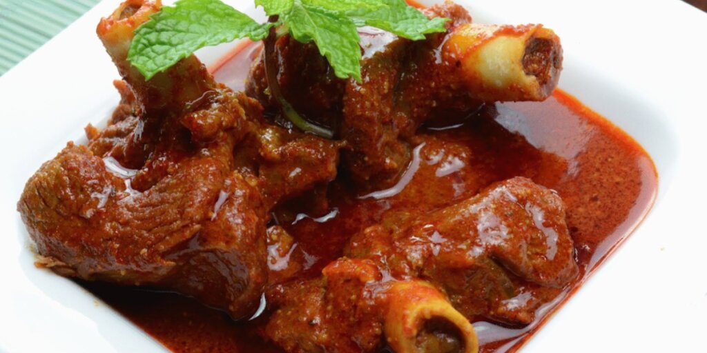 A plate of Rajasthani Laal Maas, a traditional spicy red meat curry. The dish features tender pieces of mutton coated in a rich, fiery red gravy made from red chilies and yogurt, garnished with fresh coriander leaves. The curry is served in a rustic clay bowl, accompanied by slices of fresh onions, green chilies, and lemon wedges on the side. The vibrant red color of the curry contrasts beautifully with the earthy tones of the bowl and the garnishes, evoking a sense of warmth and authenticity.