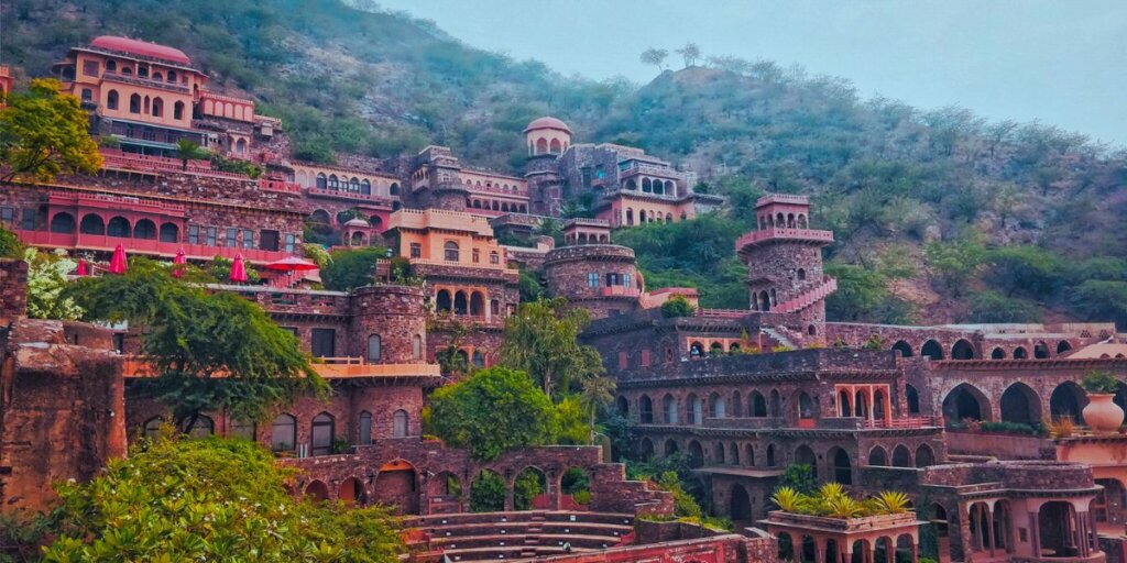 A stunning view of the Neemrana Fort in Alwar, Rajasthan, India. A historic fort built on a hill, it displays its splendid architecture with multiple levels of terraces, arched doorways, and intricate stone carvings. The fort walls and towers are surrounded by lush greenery, with a clear blue sky above, enhancing the picturesque view. The image highlights the historic grandeur and natural beauty.